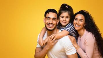 Closeup Portrait Of Cheerful Arab Family With Little Daughter Hugging And Smiling At Camera, Happy Middle Eastern Parents Bonding With Their Child While Posing Together Over Yellow Studio Background photo