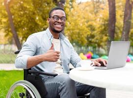 Online work for disabled people. Smiling handicapped black guy in wheelchair working on laptop, showing thumb up gesture at cafe in autumn park, recommending remote employment, copy space photo