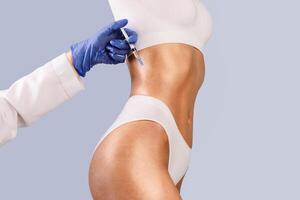 Beauty Treatment. Cropped closeup view of young fit woman getting injection in belly area, having procedure in salon. Plastic surgeon doctor in gloves making jab in stomach, holding syringe in hand photo