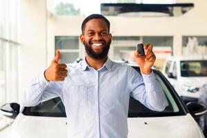 Cheerful African American Man Standing Next To His New Car, Holding Key And Showing Thumb Up. Excited Black Guy Celebrating Purchasing New Vehicle In Dealership Office, Looking And Smiling At Camera photo