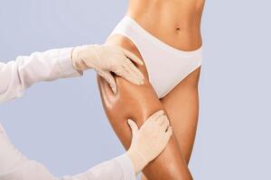 Aesthetic Medicine. Lady getting consultation at plastic surgery clinic, closeup cropped view of her legs and doctor's hands in protective gloves touching and examining thigh, making fat removal plan photo