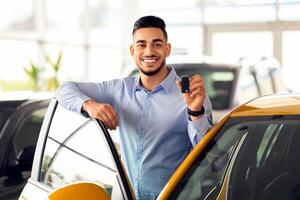 Wealthy middle-eastern businessman buying new car in luxury auto showroom, cheerful arab guy standing by nice comfy yellow automobile, showing key and smiling at camera, copy space photo
