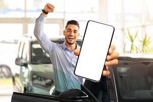 Emotional middle eastern guy choosing new car, standing by brand new black automobile, showing modern cell phone with white blank screen and raissing hand up, using auto dealership app, mockup photo