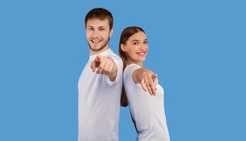 Hey, You. Portrait of cheerful casual friends pointing finger at camera, standing back to back on purple wall, copyspace photo