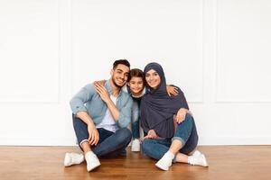 Portrait of loving happy Middle Eastern family with little preschooler kid sitting near white wall looking at camera and smiling, parents enjoy time with small child posing together in modern new home photo