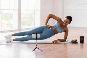 Online Workout. Smiling African Lady Standing In Side Plank Position On Yoga Mat Near Window, Looking At Mobile Phone On Tripod, Watching Tutorial Or Recording For Blog, Having Chat With Coach photo
