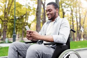 Cheery disabled black guy in wheelchair using mobile phone, checking email or messages at autumn park. Young handicapped Afro guy with cellphone posting in social network, browsing web outside photo