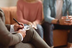 Help Concept. Closeup Cropped View Of Consultant Writing Taking Notes, Selective Focus On Hand With Pen. Spouses Having Session With Man Marriage Therapist, Sitting On Couch In Office. photo
