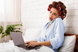 Shocked Housewife Having Issue With Laptop Computer, Talking On Cellphone Sitting In Bed At Home. Internet Connection Problem, Discontented Customer Concept. photo
