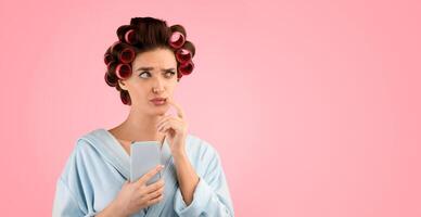 Thoughtful Glamorous Woman With Curlers Using Smartphone Looking Aside Posing Over Yellow Studio Background. Mobile Offer, New Application Concept. Panorama With Copy Space photo