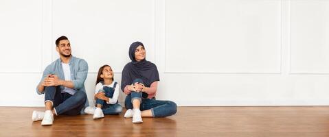 Thinking About Interior Design. Portrait of smiling muslim people sitting on floor leaning on white wall in new flat, looking aside at free copy space. Real Estate Agency Advertisement Concept photo