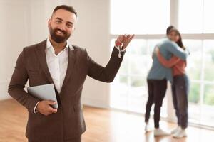 House Ownership. Portrait Of Smiling Professional Real Estate Agent In Suit Holding And Showing Key To Camera, Selective Focus. Happy Spouses Embracing Standing In New Home In Blurred Background photo