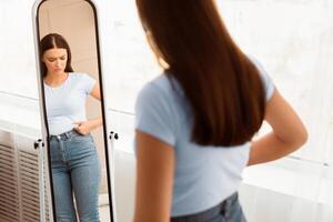 Unhappy Woman Gaining Excess Weight Touching Drooping Belly Standing At Mirror At Home. Unsuccessful Diet. Selective Focus photo