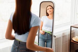Frustrated Lady Measuring Waist With Tape Gaining Excessive Weight Standing At Mirror At Home. Copy Space, Selective Focus photo