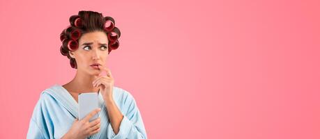Puzzled Housewife Holding Smartphone Thinking Standing On Yellow Studio Background. Pensive Lady With Hair Curlers Using Cellphone And Mobile Phone Application. Panorama, Empty Space For Text photo