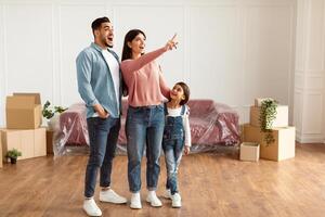 Young Homeowners Concept. Excited young family of three people planning interior, decorations and furniture in their own house, standing in empty living room, woman pointing up at free copy space photo