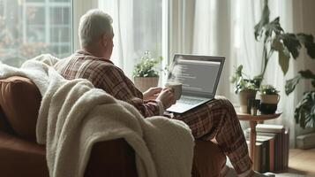 Senior man relaxing on sofa with laptop and hot drink photo