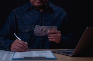 A man is sitting at a desk with a laptop and a stack of cash. He is writing something on a piece of paper photo