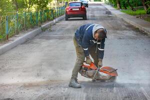 A worker cuts worn-out asphalt with a petrol cutter in a cloud of dust against the backdrop of a road being repaired. photo