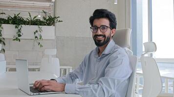 Arabian Indian muslim businessman ethnic man working with computer at corporate office cheerful happy manager employee programmer typing laptop browsing website business work looking at camera smile photo