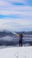 A person in a yellow jacket stands on a winter mountain summit, arms raised in joy, overlooking cloud-filled valleys and snow-covered peaks in the distance. Vertical footage video
