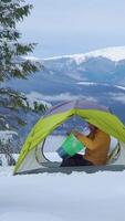 A person sits inside a tent surrounded by snow, looking at a map while enjoying a breathtaking mountain view on a chilly day. Vertical footage video