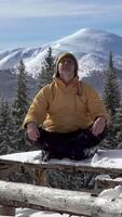 une la personne est assis jambes croisées sur une en bois Plate-forme entouré par neige, se concentrer sur méditation. majestueux montagnes monter dans le Contexte en dessous de une clair bleu ciel. verticale métrage video
