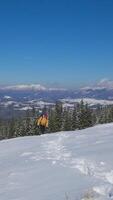 A hiker dressed in bright yellow weather gear walks through fresh snow, carrying a backpack. The stunning winter landscape features snow-capped mountains. Vertical foot video