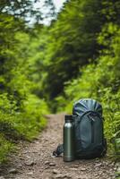 On a dirt trail, a sleek water bottle and a compact backpack rest amidst lush greenery. The setting promotes adventure and exploration, inviting outdoor enthusiasts to enjoy nature photo
