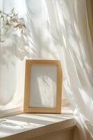 A light oak picture frame rests on a windowsill, illuminated by soft natural light streaming through sheer white curtains, evoking a peaceful, airy ambiance perfect for displaying memories photo