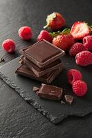 Delicious dark chocolate squares are stacked elegantly on a textured black slate. Surrounding them are fresh raspberries and strawberries, creating a vibrant and inviting display photo