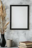 A sleek picture frame with a thin black border is mounted on a textured concrete wall. Nearby, a ceramic vase holds dried grass, and a stack of books adds a touch of minimalistic style photo