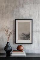 A sleek picture frame with a thin black border hangs against a textured concrete wall. It is accompanied by a ceramic vase and a neatly stacked set of books, creating a chic minimalist aesthetic photo