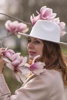 Woman magnolia flowers, surrounded by blossoming trees., hair down, white hat, wearing a light coat. Captured during spring, showcasing natural beauty and seasonal change. photo