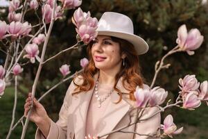 Woman magnolia flowers, surrounded by blossoming trees., hair down, white hat, wearing a light coat. Captured during spring, showcasing natural beauty and seasonal change. photo