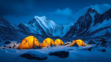 Illuminated orange tents glow against snowy mountain landscape with dramatic snow-capped peaks, creating a moody base camp setting in high-altitude Himalayan-style terrain during blue evening light H photo