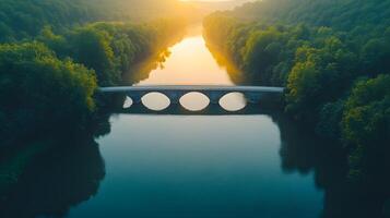 Modern bridge spans tranquil river during golden sunset, surrounded by lush forests with perfect water reflections, captured from drone perspective in misty evening atmosphere. sunset photo