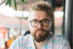 Close-up portrait of a man with glasses, curly hair, and beard, sitting outdoors at a cafe. Neutral expression and natural lighting highlight individuality and a calm, thoughtful demeanor. Millennial photo