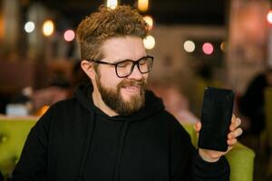 Satisfied man with dark beard holding smartphone and showing his favorite mobile app Male programmer has developed a cool application. photo