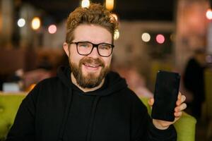Satisfied man with dark beard holding smartphone and showing his favorite mobile app Male programmer has developed a cool application. photo