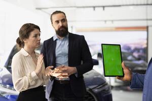 Car sales executive showcasing dealership vehicles to client using green screen tablet. Showroom salesperson helping customer choose automobile using chroma key device, comparing models photo
