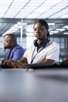 African american engineer in data center using diagnostic tools and system monitoring software on computers to identify errors. Woman next to colleague in server farm repairing infrastructure photo