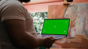 African american artist holding device with green screen mockup in art studio, working on a new painting with a certain technique. Painter using watercolor and acrylic dye for his creation. Camera B. photo