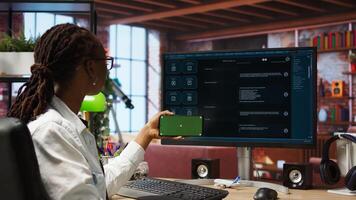 Woman seated at desk using isolated screen mobile phone to follow tutorial on AI LLM chatbot software. African american person uses mockup smartphone in apartment and GPT program on computer, camera A photo