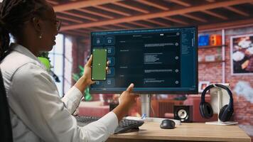 Woman seated at desk using AI LLM chatbot software and green screen phone in modern urban home office. Person uses large language model program on computer and mockup smartphone in apartment, camera B photo