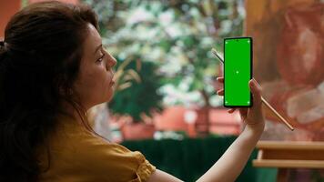 Young woman looking at chroma key display and at her rustic masterpiece, feeling proud and confident with her paintwork for the art gallery. Artist enjoying her hobby for leisure. Camera A. photo