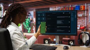 Woman seated at desk using AI LLM chatbot software and green screen phone in modern urban home office. Person uses large language model program on computer and mockup smartphone in apartment, camera A photo
