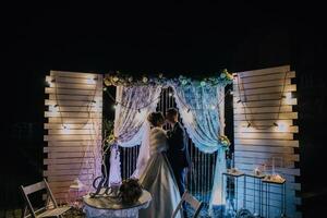 A bride and groom are standing in front of a white archway with a sign that says photo