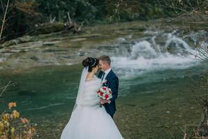 A bride and groom are standing in front of a waterfall, holding hands photo