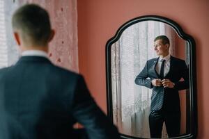 A man in a suit is standing in front of a mirror, adjusting his tie. photo